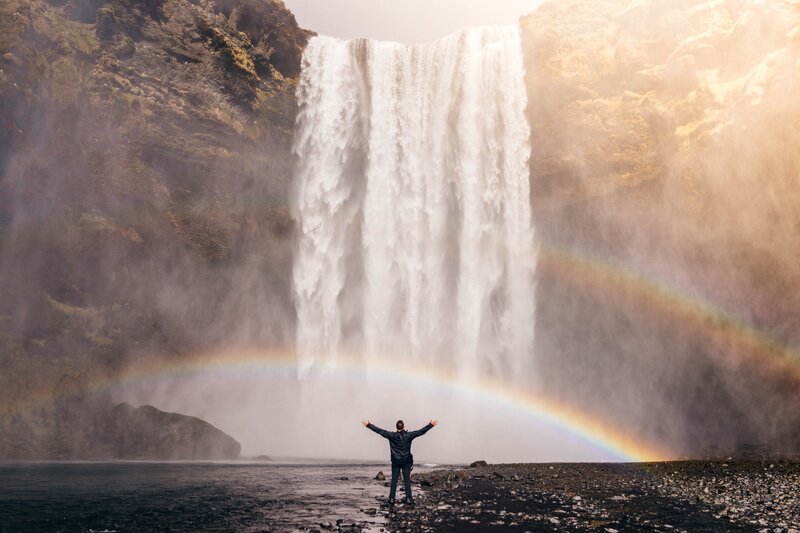 Image of a man standing with his arms outstretched underneath a rainbow and waterfall