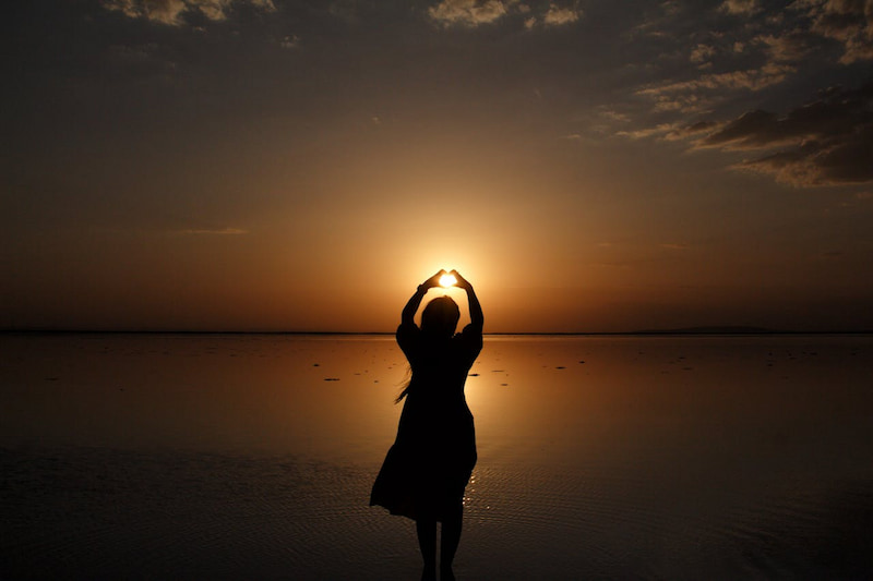 Image of a person in front of a calm ocean at sunset with her hands held up to form a love heart